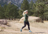 Woman running by large rocks and trees near mountains, wearing Aqua X Sport runners