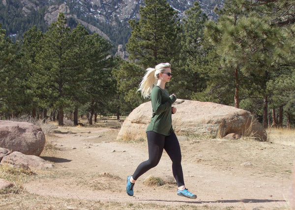 Woman running by large rocks and trees near mountains, wearing Aqua X Sport runners