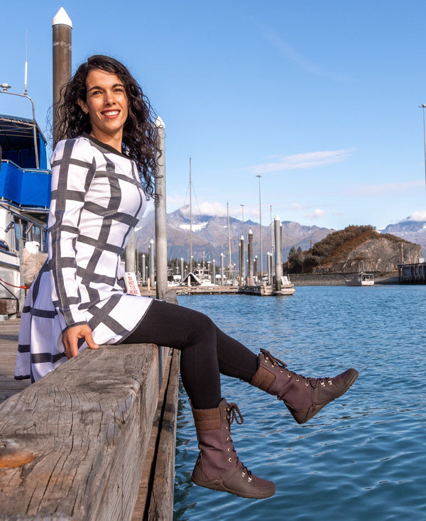 Sitting on the edge of a high dock wearing Plum Mika at the water, black leggings and a white and black tunic